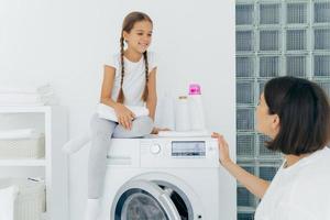 schattig klein meisje met staartjes, poseert bovenop de wasmachine, houdt een witte zachte handdoek vast, kijkt blij naar moeder, praat over plannen na het wassen. brunette huisvrouw laadt wasmachine, bezig zijn foto