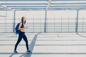 buitenopname van mooie vrouw met slank lichaam, gekleed in casual top en legging, draagtas, gaat naar de sportschool, gebruikt moderne smartphone om naar muziek te luisteren, kopieer ruimte opzij voor uw advertentie foto