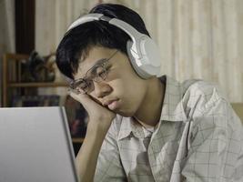 verveelde slaperige student die een koptelefoon draagt, moe van het leren huiswerk maken, half slapend op het laptopscherm in zijn kamer. triest tienerjongen studeren voorbereiding voor examen test. foto