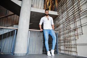 stijlvolle lange Arabische man model in wit overhemd, jeans poseerde tegen stalen muur binnen. baard aantrekkelijke Arabische man. foto