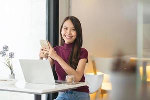 jonge aziatische vrouw die telefoon en laptop in coffeeshop gebruikt en naar camera kijkt. foto