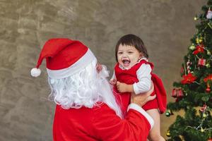 De kerstman tilt een gelukkig klein peutermeisje op en lacht vrolijk met een volledig versierde kerstboom op de achterkant voor het concept van de seizoensviering foto