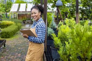 Afro-Amerikaanse tuinman die klembord vasthoudt terwijl hij in haar coniferenboomkwekerij werkt voor het leveringsconcept van groenblijvende en bonsaikunstenaars foto