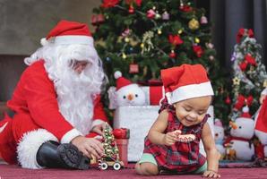 afro-amerikaanse baby heeft plezier met spelen met speelgoed terwijl de kerstman een cadeau voorbereidt en achter de kerstboom zit voor het concept van de seizoensviering foto