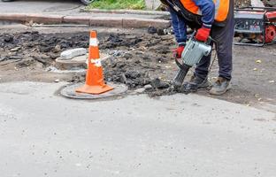 een arbeider met een elektrische drilboor breekt het oude asfalt van de weg rond het rioolmangat. foto