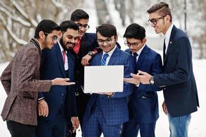 groep van zes Indiase zakenman in pakken poseerde buiten in de winterdag in Europa, kijkend op laptop. foto