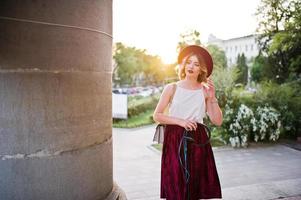 modieuze en mooie blonde model meisje in stijlvolle rood fluwelen velours rok, witte blouse en hoed, poseerde met telefoon en koptelefoon tegen kolom bij zonsondergang. foto