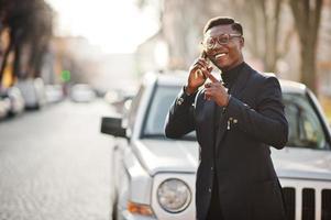 verbazingwekkend uitziende Afro-Amerikaanse man draagt een blauwe blazer met broche, zwarte coltrui en een bril op straat. modieuze zwarte man die telefonisch tegen auto spreekt. foto