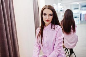 brunette prachtig meisje in de kledingwinkel boetiek bij casual kleding en roze blouse tegen spiegel. foto