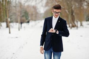 stijlvolle Indiase student man in pak en bril poseerde op winterdag buiten, kijkend naar zijn horloges. foto