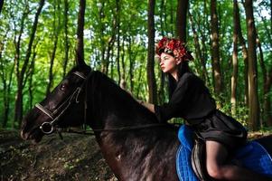 mystieke meisje in krans slijtage in het zwart bij paard in hout. foto