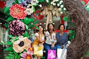 mooie drie goed geklede afro-amerikaanse meisjes met gekleurde boodschappentassen zittend op de fotozone van de lentedecoratie in het winkelcentrum. foto