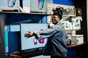 stijlvolle casual Afro-Amerikaanse man bij jeansjasje en zwarte baret in de buurt van nieuw tv-scherm bij elektronicawinkel. foto