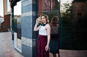 modieuze en mooie blonde model meisje in stijlvolle rood fluwelen velours rok, witte blouse, geposeerd met telefoon en koptelefoon tegen ramen van stadsgebouw. foto