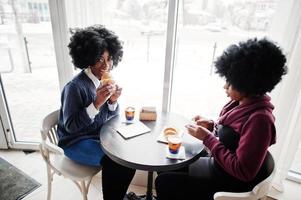 twee Afro-Amerikaanse vrouwen met krullend haar dragen op truien zit aan tafelcafé, eet een croissant en drinkt thee. foto