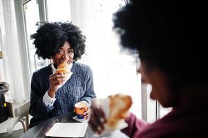 twee Afro-Amerikaanse vrouwen met krullend haar dragen op truien zit aan tafelcafé, eet een croissant en drinkt thee. foto