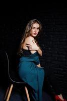 knappe brunette meisje slijtage op zwarte en groene jas in rode hoge hakken, zittend en poseren op stoel in studio tegen donkere bakstenen muur. studiomodel portret. foto