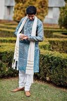 indiase man draagt traditionele kleding met witte sjaal buiten tegen groene struiken in het park, kijkend naar zijn horloges. foto