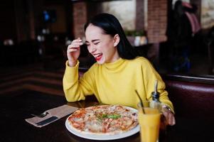 grappige brunette meisje in gele trui pizza eten in restaurant. foto