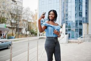 aantrekkelijke afro-amerikaanse vrouw met dreadlocks in spijkerbroek, geposeerd in de buurt van balustrades tegen modern gebouw met meerdere verdiepingen dat selfie maakt op mobiele telefoon. foto