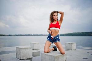 sexy krullend model meisje in rode top, jeans denim shorts en sneakers geposeerd op stenen kubussen tegen bewolkte hemel en meer. foto