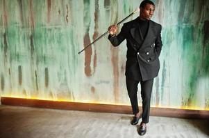 stijlvolle afro-amerikaanse heer in elegante zwarte jas, met retro wandelstok als rieten kolf of kantelstok met gouden diamanten balhandvat. rijke modieuze afro man tegen groene muur. foto