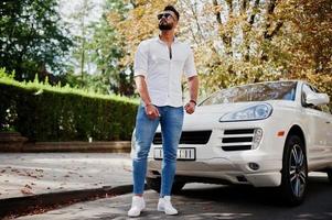 stijlvolle lange Arabische man model in wit overhemd, jeans en zonnebril gesteld op straat van de stad. baard rijke aantrekkelijke arabische man tegen witte suv auto. foto