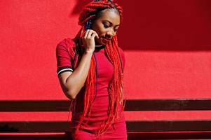 schattig en slank Afrikaans Amerikaans meisje in een rode jurk met dreadlocks buiten tegen de rode muur. stijlvol zwart model. foto