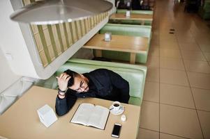 arabische man draagt een zwarte spijkerjas en een bril die in café zit, boek leest en koffie drinkt. stijlvolle en modieuze Arabische modelman. foto