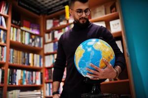 lange slimme arabische student man, draag violet coltrui en bril, in bibliotheek met earth globe bij de hand. foto