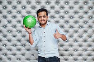 stijlvolle aziatische man in jeans shirt staande met bowlingbal bij de hand tegen zilveren muur achtergrond en duim opdagen. foto