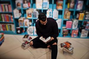 lange slimme arabische student man, draag violet coltrui en bril, in bibliotheek zittend tegen boekenplanken. foto