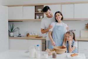 gelukkige familie koken samen in de keuken. vader, moeder en dochter druk bezig met het bereiden van een heerlijke maaltijd thuis. man omhelst vrouw die zwaait en deeg bereidt, koekjes bakt. eten, samenzijn foto