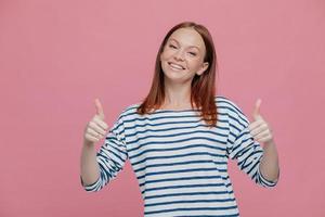 taille shot van vrij tevreden jonge vrouw draagt gestreepte trui, steekt twee duimen op, toont goed gebaar en instemming, glimlacht breed, geïsoleerd over roze achtergrond. lichaamstaal concept foto