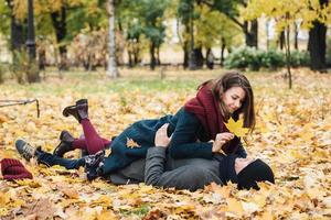 verliefd paar hebben samen plezier, liggen op de grond bedekt met gele bladeren, kijken elkaar aan, spelen dwazen. mooie vrouw draagt jas en sjaal ligt op vriendje, laat hem geel blad zien foto