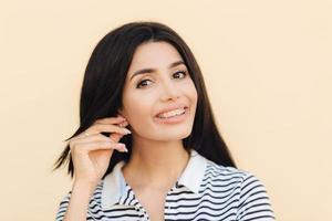 headshot van schattige mooie vrouw heeft een brede glimlach, donker haar, houdt de hand op het oor, toont witte tanden met beugels, kijkt direct in de camera, heeft een gelukkige uitdrukking, geïsoleerd over studio-achtergrond. foto