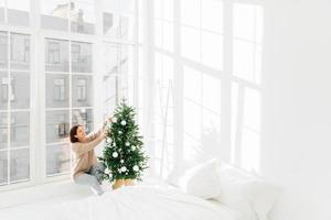 wintervakantie, nieuwjaarsconcept. blije jonge brunette vrouw heeft een feestelijke stemming, versiert de kerstboom thuis, bereidt zich voor op het feest, poseert in een gezellige slaapkamer met witte muren op de vensterbank. foto