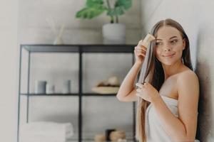 aantrekkelijke jonge vrouw glimlacht en geniet van haar schoonheid, kamt lang steil haar na het aanbrengen van een masker, staat gewikkeld in een badhanddoek in een gezellige badkamer, heeft een gladde huid. haarverzorging, spa en beauty concept foto