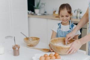 glimlachende kleine meidhelper houdt grote kom vast, kijkt hoe moeder eieren met bloem mengt, bereidt smakelijke cake met chocolade, poseert tegen een gezellig keukeninterieur, bereidt voedsel voor het hele gezin foto