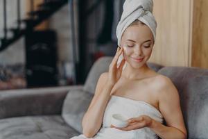 mooi vrouwelijk model geniet van zachtheid van de huid na het nemen van een bad, brengt vochtinbrengende crème op de wang aan, draagt minimale make-up, gewikkeld in een handdoek, zit alleen op de bank, ondergaat schoonheidsbehandelingen foto