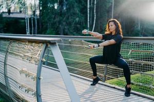 gemotiveerde brunette actieve jonge vrouw doet squat oefeningen met halters, traint biceps, gekleed in zwarte actieve kleding, poseert bij brug buiten tijdens zonsopgang, heeft ochtendtraining. sport concept foto