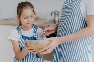 druk meisje houdt kom vast, kijkt hoe moeder ingrediënten klopt, leert koken. onherkenbare moeder in gestreepte schort brengt tijd door met dochter, laat zien hoe deeg te bereiden. kooktijd foto