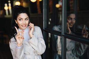 charmante jonge brunette vrouw met aangename glimlach met zachte handen en mooie manicure die via de mobiele telefoon communiceert met haar vriend die haar vinger opsteekt terwijl ze een goed idee krijgt voor een feestje foto