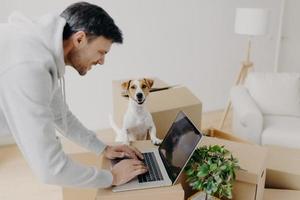 tevreden hardwerkende man druk bezig met het zoeken naar informatie op een laptopcomputer die op kartonnen dozen staat, zijn huisdier poseert in de buurt, verhuist naar een nieuw appartement, een bank en een staande lamp. onroerend goed concept foto