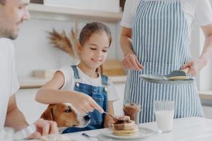 blij klein meisje geniet van het eten van een smakelijk dessert bereid door mama, voegt gesmolten chocolade toe aan pannenkoeken, geniet van het samenzijn en moeder, vader en hond, heeft een heerlijk voedzaam ontbijt in de keuken foto