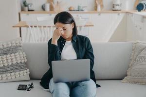 overwerkte vrouw lijdt aan vermoeidheid van de ogen, hoofdpijn, moe van het werken op een laptop die thuis op de bank zit foto