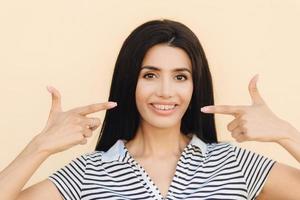 portret van aangenaam ogende brunette vrouw heeft een aantrekkelijke uitstraling, geeft aan met beide voorvingers naar de mond een zachte glimlach te tonen, tanden met beugels, gekleed in vrijetijdskleding, geïsoleerd op een lichte muur foto