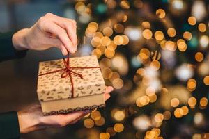 onherkenbare vrouw wikkelt geschenkdoos over kerstboom met glanzende lichten en slingers. versierde huidige doos in handen van de vrouw. vakantie en viering concept. foto