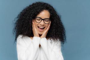 overgevoelige vrouw raakt wangen aan lacht van vreugde, houdt ogen gesloten, kan niet stoppen met giechelen, heeft afro-kapsel, gekleed in witte trui, modellen tegen blauwe achtergrond. etniciteit en emoties concept foto