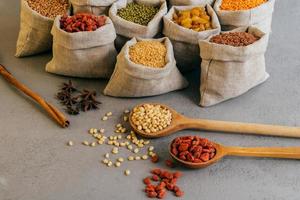 shot van kleine zakjes met kleurrijke granen, voedzame peulvruchten, steranijs in de buurt, twee houten lepels met rode goji-bessen. rauwe producten foto